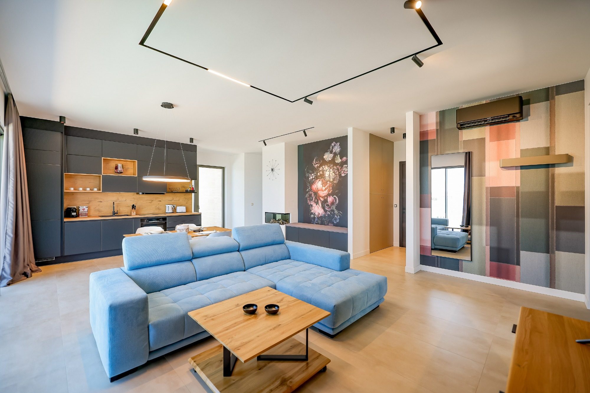 Moderne Wohnzimmer mit blauer Couch, Holztisch und offener Küchenzeile im Hintergrund.
